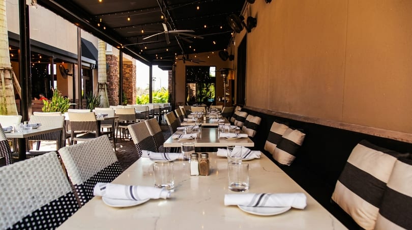 An empty outdoor patio dining area with set tables, black and white striped cushions, and string lights.