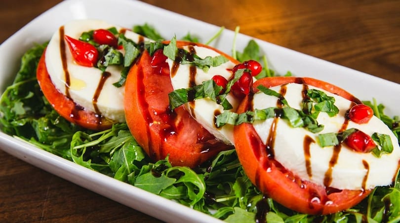 Three Caprese salad servings with tomatoes, mozzarella, basil, and balsamic glaze on a bed of arugula.