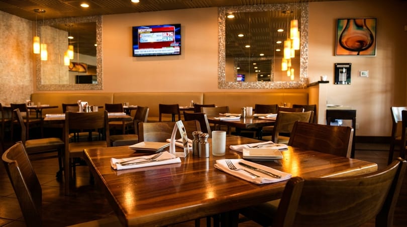 Warmly lit restaurant interior with wooden tables set with white napkins and cutlery, and a TV on the wall.